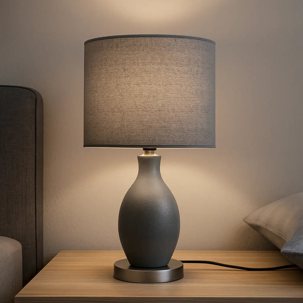 Lampe de chevet grise moderne avec abat-jour cylindrique sur une table en bois clair dans une chambre épurée.